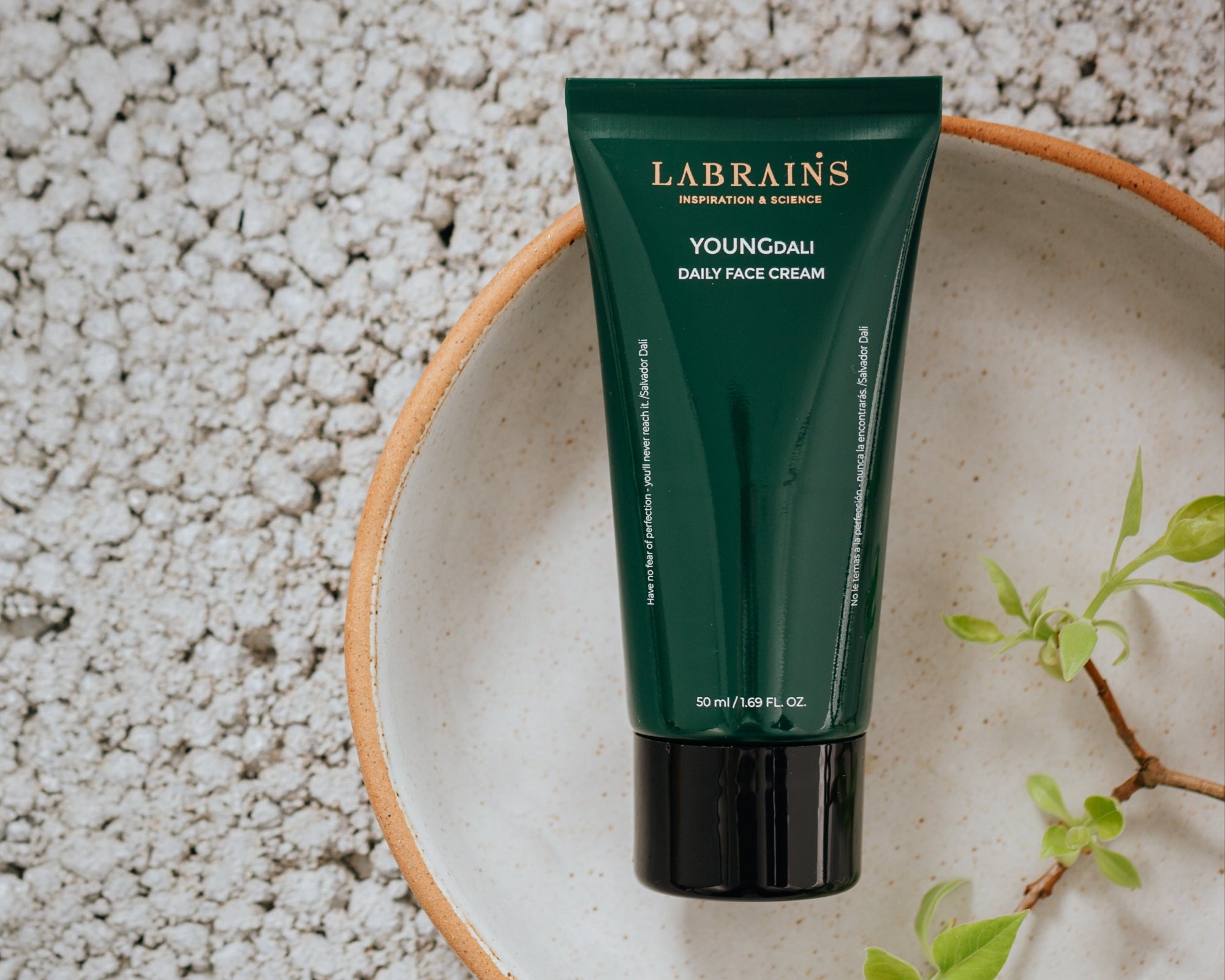 Green tube of Labrains YoungDali Daily Face Cream on a textured surface with a plant branch.