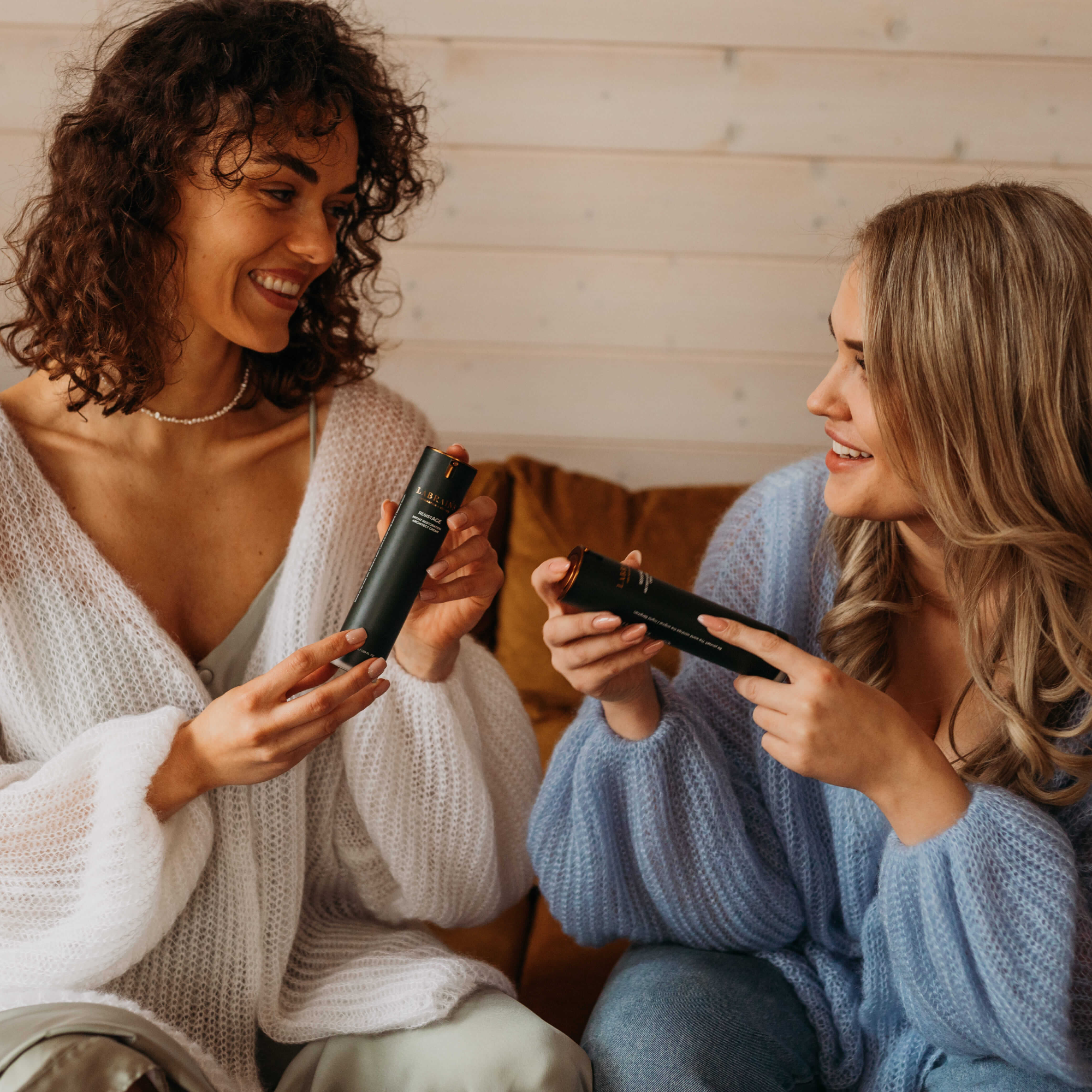 Two women sitting on a couch, holding labrains skincare products and smiling.