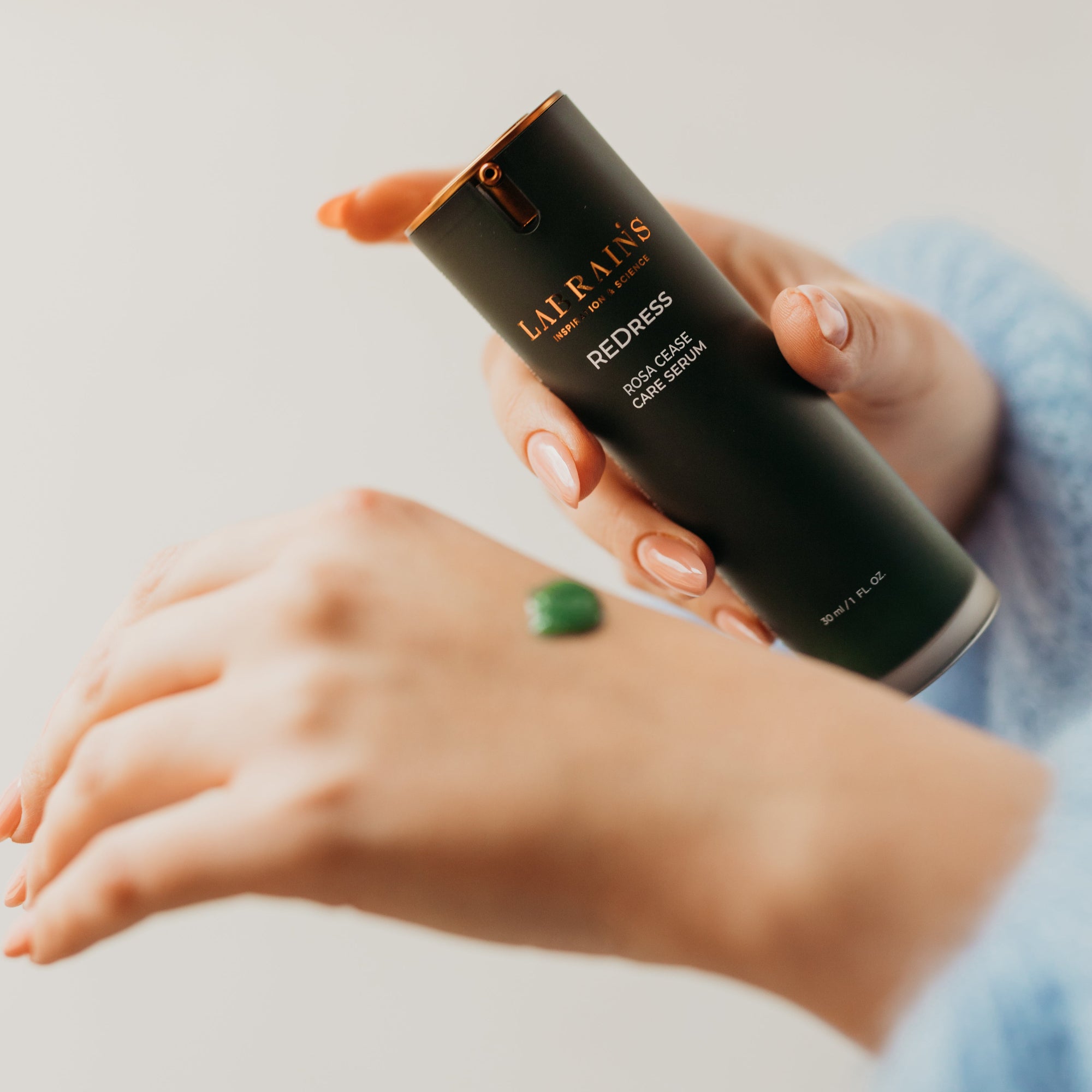 Woman's hands holding a labrains skincare product labeled 'redress rosa cease care serum' on a neutral background