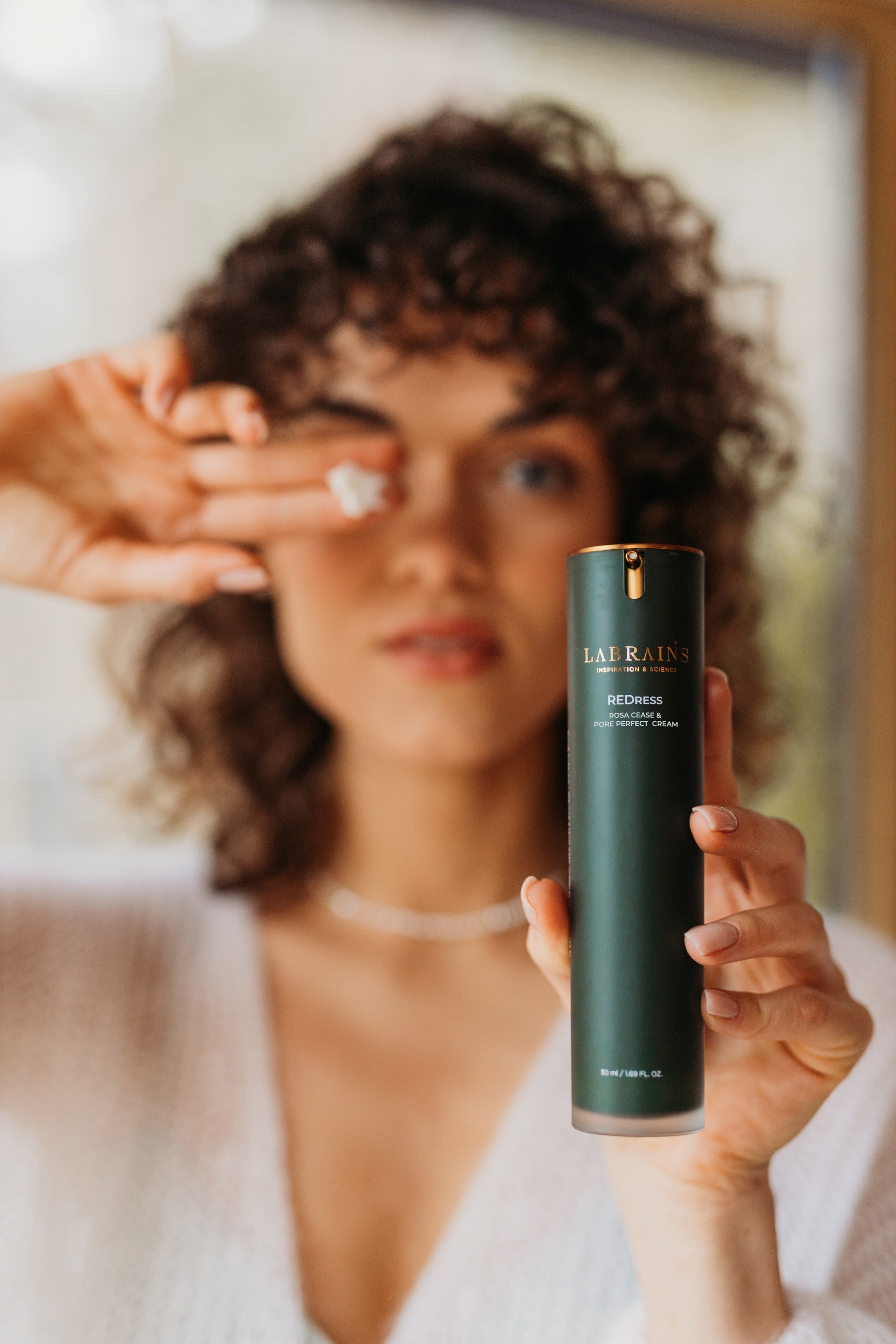 curly woman holding a green bottle of labrains skincare product labeled 'redress rosa cease & strike back cream' in front of her face